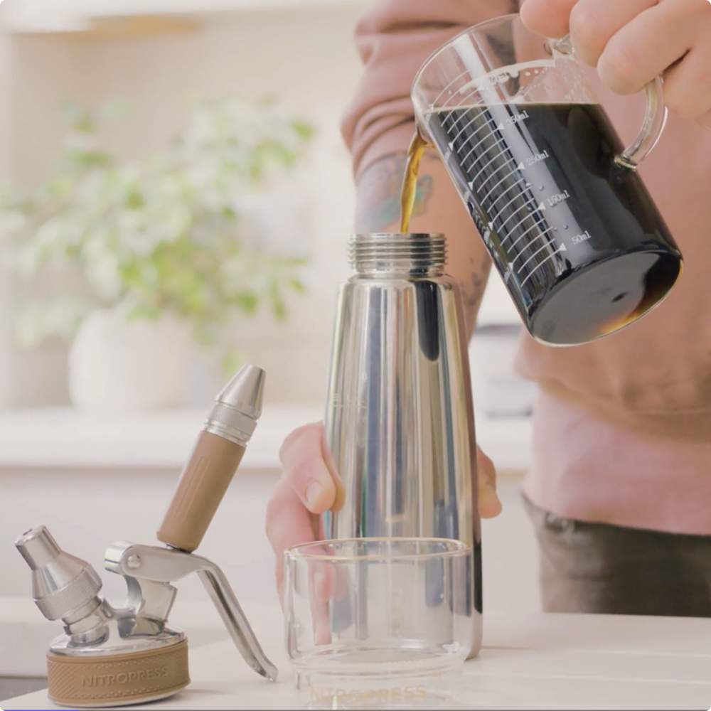 Person pouring cold brew from a glass container into a nitropress with a blurred background